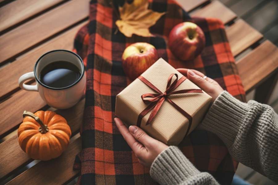 Mains tenant un cadeau Halloween kraft avec ruban bordeaux, entouré d'une citrouille, d'un café et de pommes sur une table en bois automnal