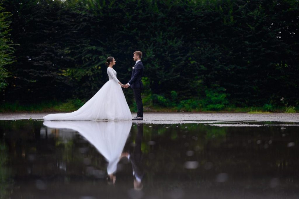 Mariés se tenant la main face à face avec reflet dans une flaque d'eau, illustrant des idées photos de mariage sous la pluie.